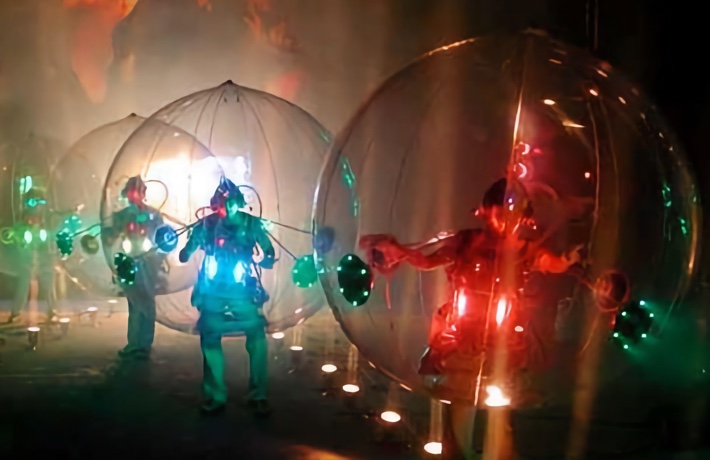 Des personnes enfermées dans d’immenses bulles transparentes défilent sous des projecteurs.