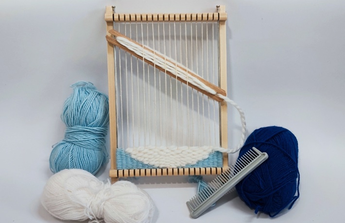 Petite planche à tisser avec des pelotes de laine bleu et blanches à côté, et le début indiscernable d’un tissage.