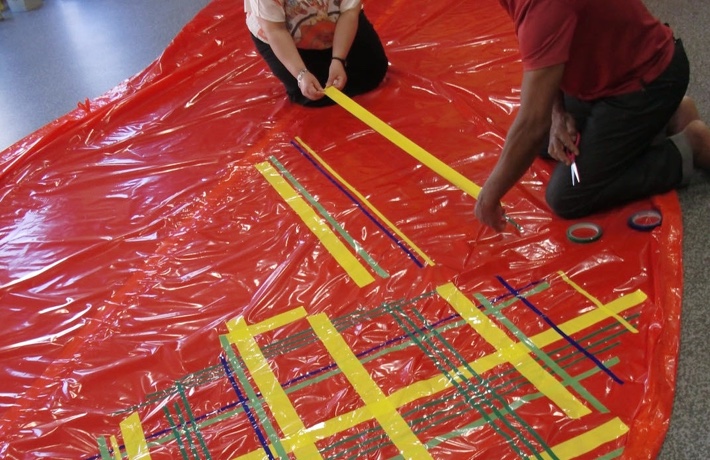 Deux personnes posent un motif de lignes à base de rubans de différentes couleurs sur une grande bâche rouge.