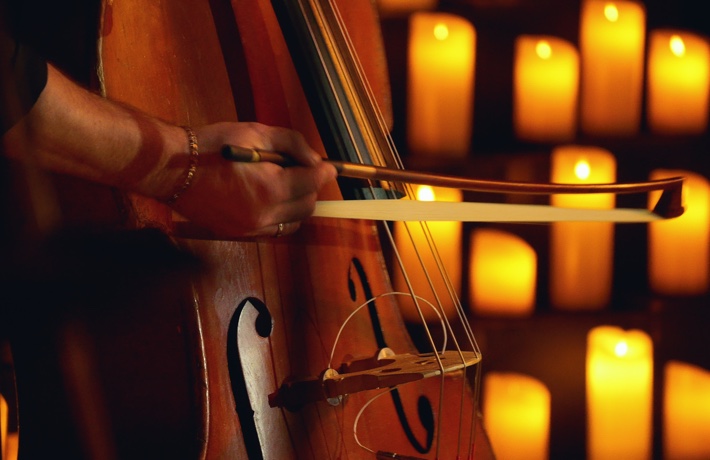 Gros plan sur l’arche qu’une personne pose sur un violoncelle, avec des bougies en arrière-plan.