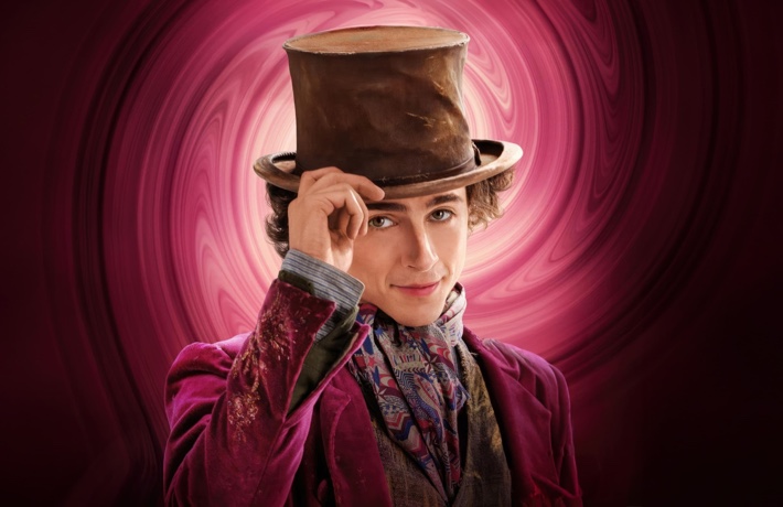 Timothée Chalamet dans Wonka, s’apprête à soulever son chapeau d’un air espiègle, avec un arrière-plan rouge-rose tourbillonnant.