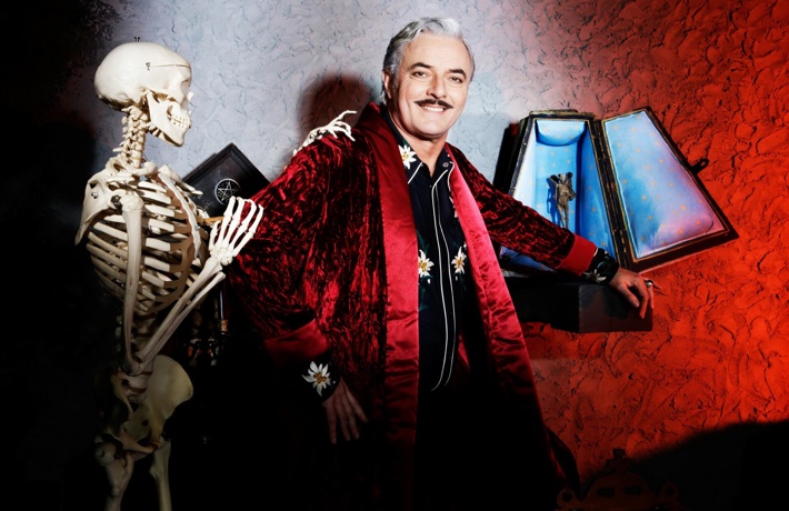 Un magicien sourit à l’objectif. Un squelette en taille réelle est tourné vers lui et a sa main posée sur l’épaule du magicien.