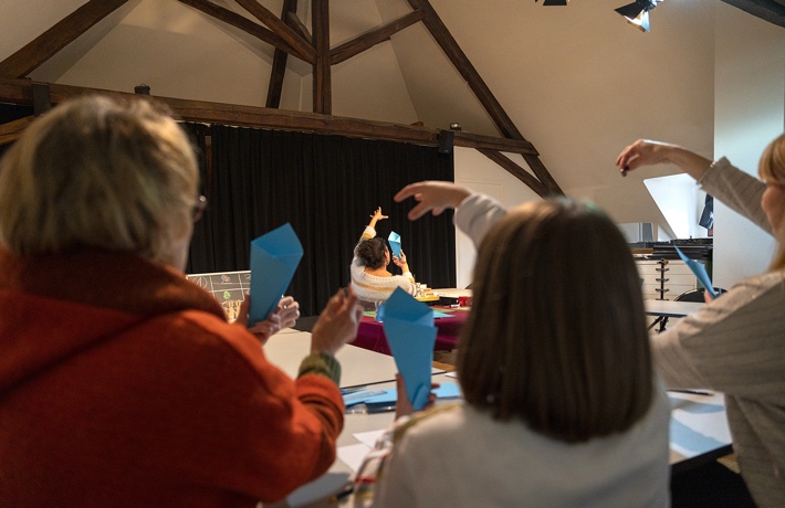 Dans une grande salle, des personnes s’exercent à faire un tour de magie faisant tomber une bille dans une feuille de papier tenue en cylindre, suivant l’exemple d’une magicienne à l’arrière-plan.