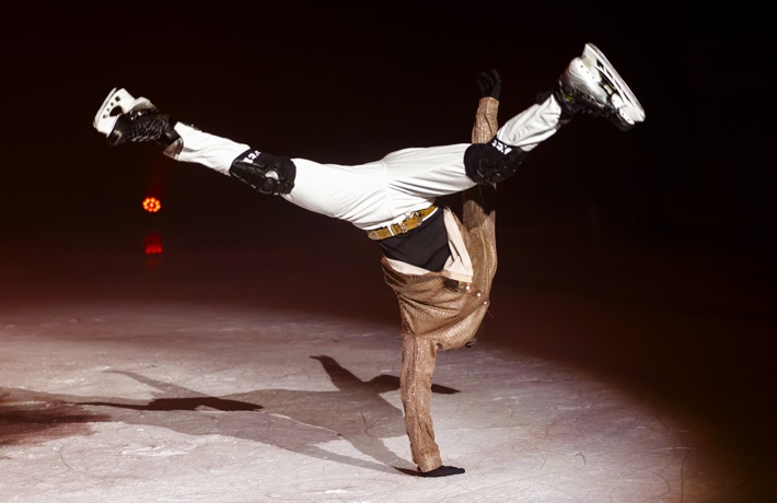 Une personne fait une figure de danse en patins sur la glace : elle est complètement renversée, la main sur la glace, les pieds en l’air.