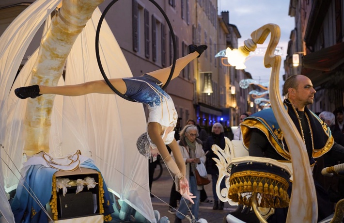 Une personne danse de façon acrobatique, totalement retournée dans un cerceau, avançant dans un char. Devant, un musicien en costume.