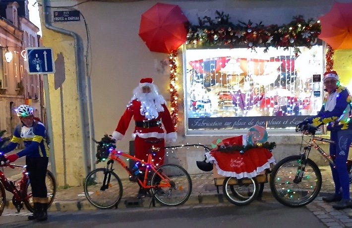 Trois cyclistes, dont un déguisé en père Noël et ayant une remorque accrochée à son vélo. Chaque vélo est recouvert d’une guirlande lumineuse.